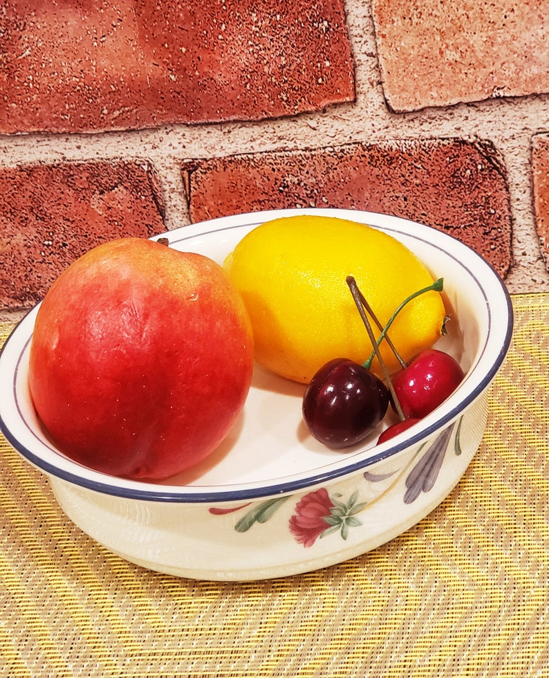 May include: A white ceramic bowl with a blue rim and floral design, containing a red peach, a yellow lemon, and two red cherries.