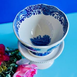 May include: A white porcelain teacup and saucer set with a blue floral and landscape design. The teacup has a scalloped rim and sits atop a pedestal base. The design includes a castle scene.