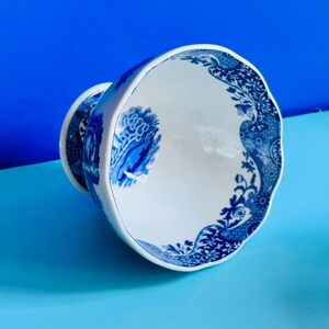 May include: White and blue ceramic teacup with a decorative floral and landscape design. The teacup has a scalloped rim and a matching blue and white design on the base. The teacup is set against a blue background.