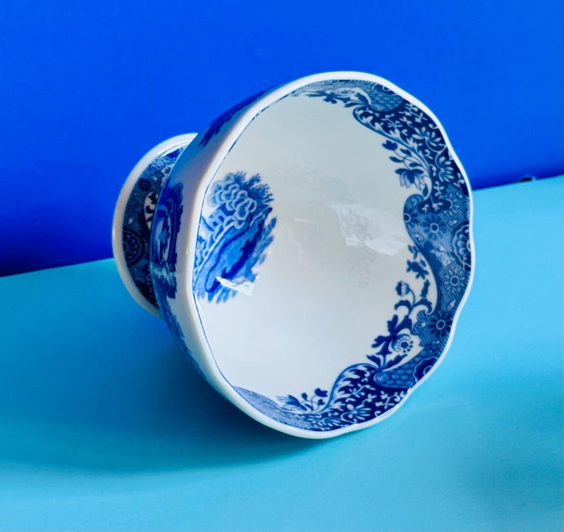 May include: White and blue ceramic teacup with a decorative floral and landscape design. The teacup has a scalloped rim and a matching blue and white design on the base. The teacup is set against a blue background.