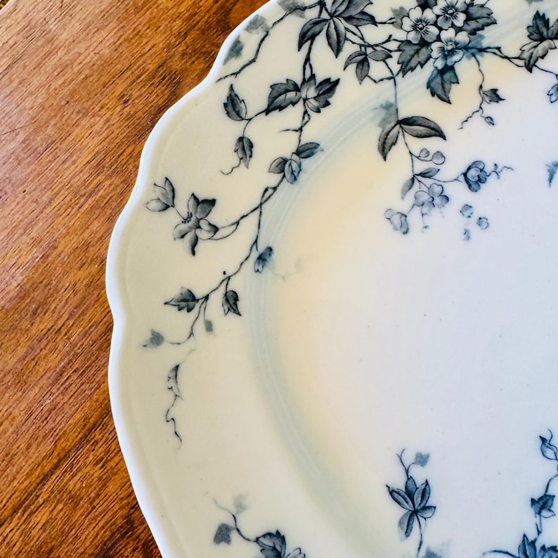 May include: A white ceramic plate with a scalloped edge, adorned with a delicate floral pattern in shades of blue and gray. The design features trailing vines and clusters of flowers, creating an elegant and vintage aesthetic. The plate is set against a warm wooden surface.