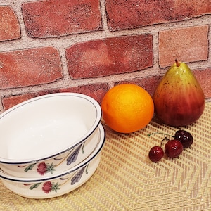 May include: Two white ceramic bowls with a floral design, stacked on top of each other. The bowls are on a woven placemat with a pear, orange, and cherries.