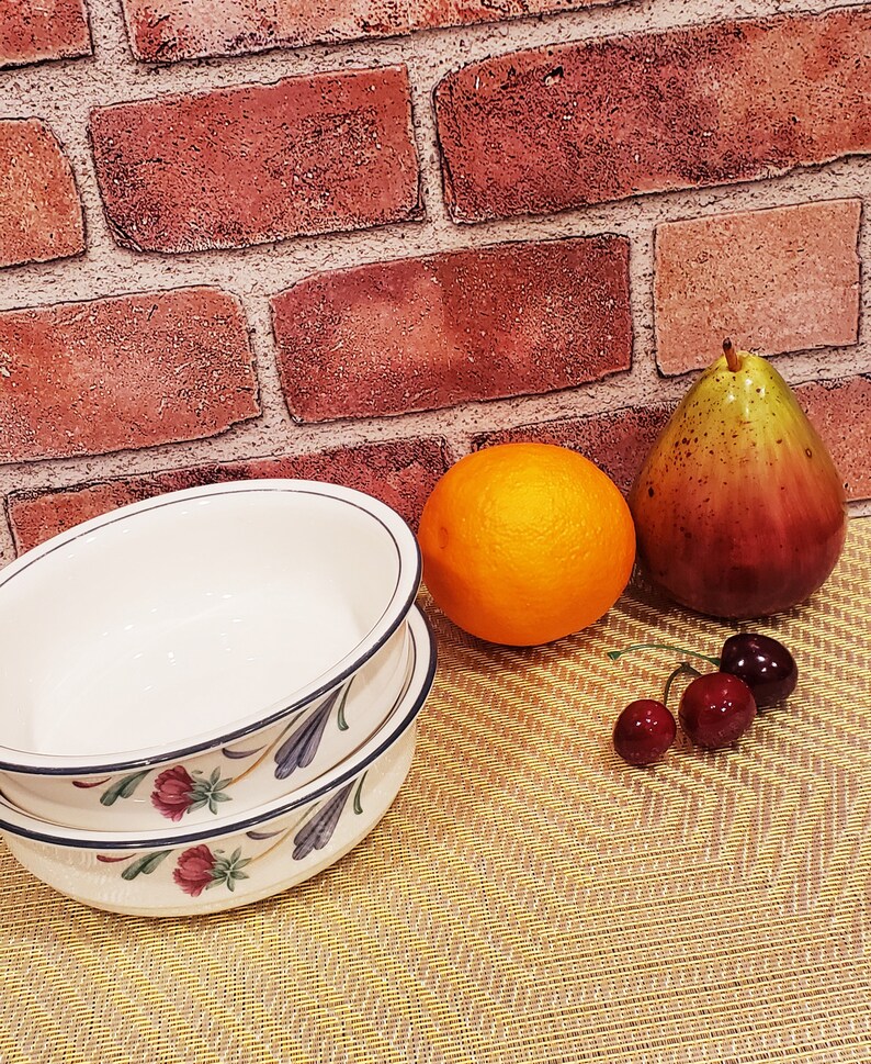 May include: Two white ceramic bowls with a floral design, stacked on top of each other. The bowls are on a woven placemat with a pear, orange, and cherries.