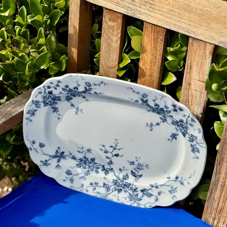 May include: A white, oval serving platter with a scalloped edge. The platter is decorated with a blue floral pattern. The design features clusters of flowers and trailing vines. The platter is resting on a blue surface and wooden slats.