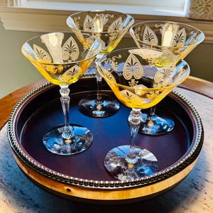 May include: A set of four yellow glass cocktail glasses with clear stems and bases. Each glass features a white etched pattern of stylized spade shapes and scrolling flourishes around the rim. The glasses are arranged on a round dark tray.