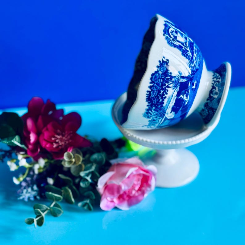 May include: A white and blue porcelain bowl with a pedestal base. The bowl features a blue landscape design. The image also includes pink and red flowers and greenery, all set against a blue background.