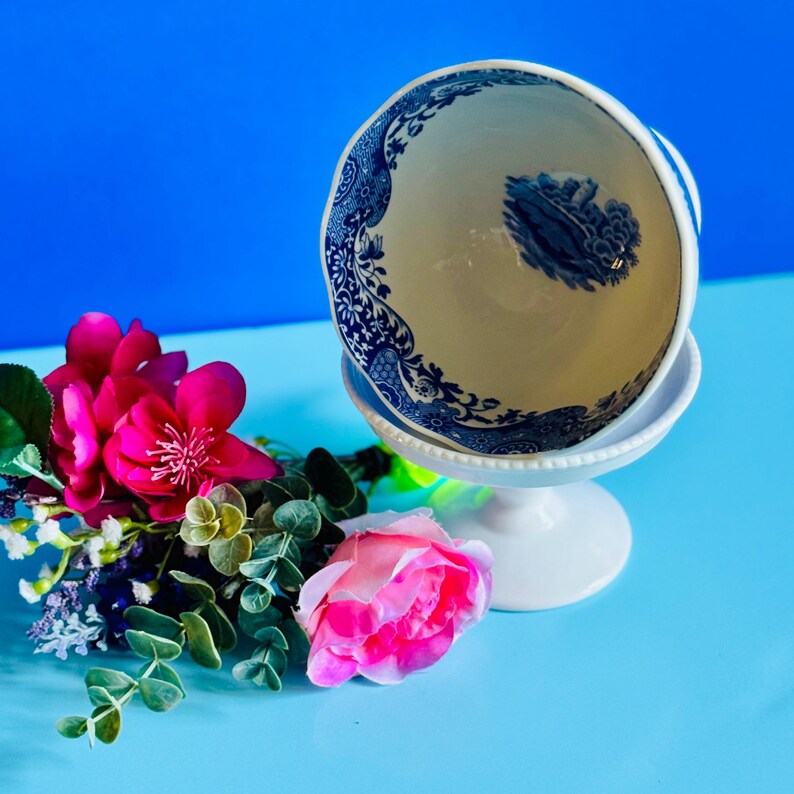 May include: A white pedestal bowl with a blue and white floral design. The bowl is filled with a pink flower arrangement. The background is a bright blue.