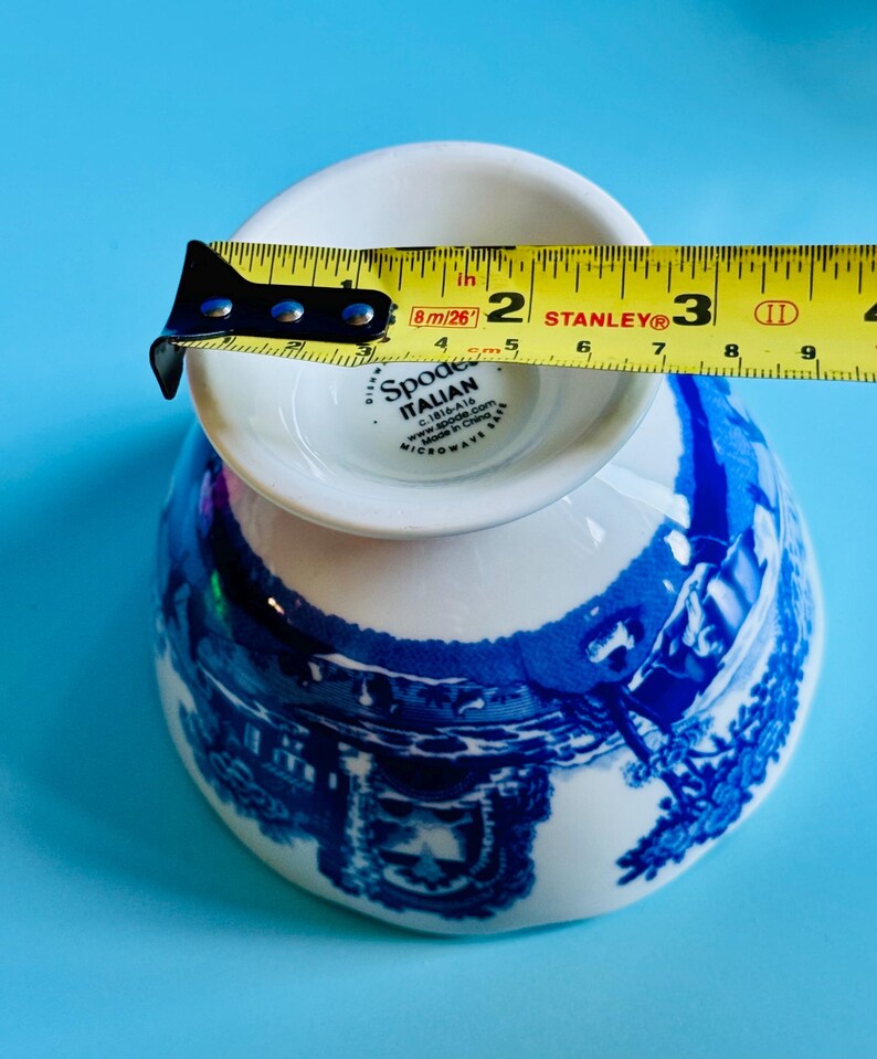 May include: A white and blue Spode Italian teacup, with a blue scenic design. The teacup is measured with a yellow tape measure, showing a diameter of approximately 2 inches.