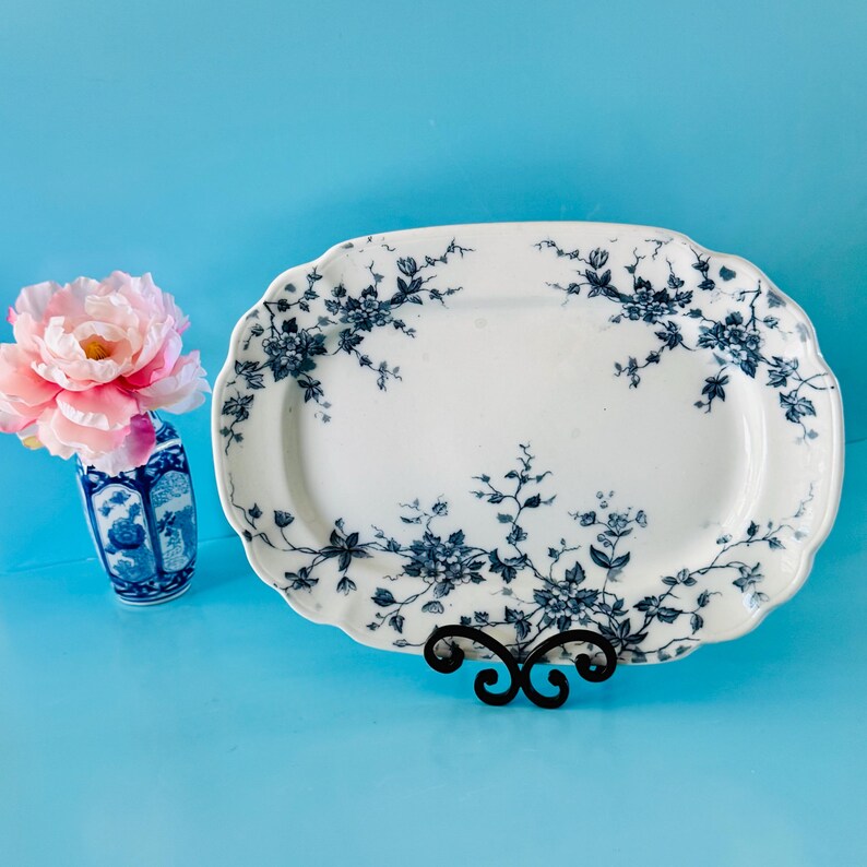 May include: White oval serving platter with blue floral design, displayed on a black metal stand. A small blue and white vase holds pink flowers, set against a blue background.