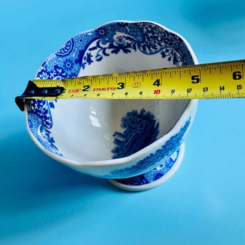May include: A white and blue porcelain bowl with a scalloped rim and pedestal base. The bowl features a blue floral pattern and measures approximately 5 inches in diameter. The bowl is set against a blue background.