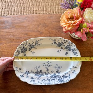 May include: An oval, white serving platter with a blue floral design. The platter measures approximately 15 inches long. The edges are scalloped. A bouquet of flowers is in the background.