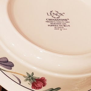 May include: A white ceramic bowl with a floral design. The bowl is made by Lenox and is microwave safe. The text on the bottom of the bowl reads "LENOX&reg; CHINASTONE&reg; FREEZER TO OVEN TO TABLE MICROWAVE SAFE POPPIES ON BLUE &copy;LENOX MADE IN USA"