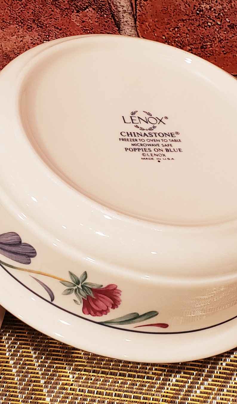 May include: A white ceramic bowl with a floral design. The bowl is made by Lenox and is microwave safe. The text on the bottom of the bowl reads "LENOX&reg; CHINASTONE&reg; FREEZER TO OVEN TO TABLE MICROWAVE SAFE POPPIES ON BLUE &copy;LENOX MADE IN USA"