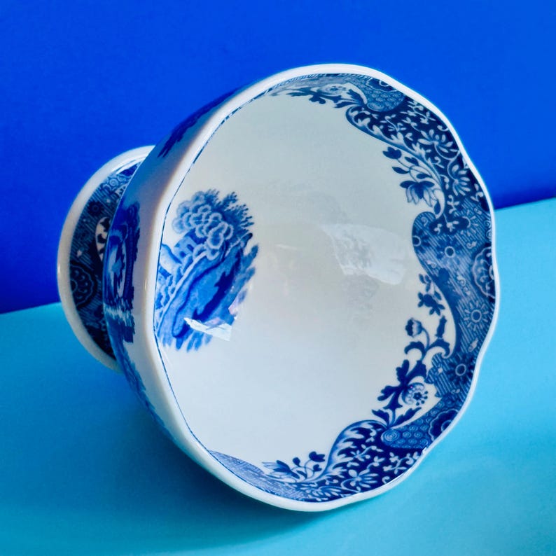 May include: A white teacup with a blue floral design. The cup has a scalloped edge and a handle. The blue pattern includes floral and landscape motifs. The teacup is set against a blue background.