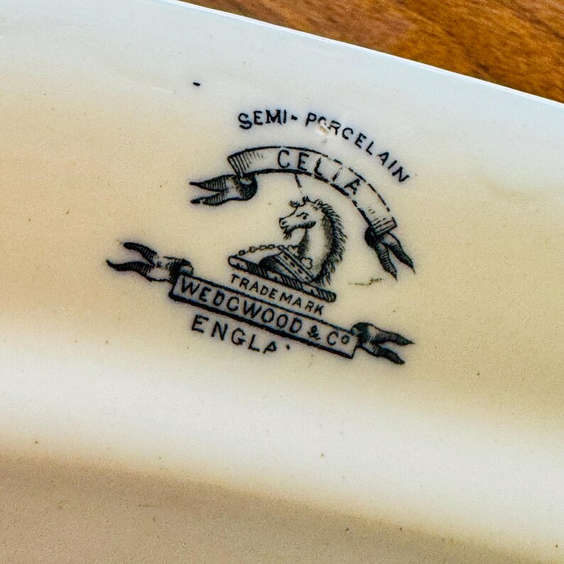 May include: Close-up of a white ceramic dish with a black and white emblem. The emblem features a horse and the words "SEMI-PORCELAIN CELIA" and "WEDGWOOD & CO ENGLA".