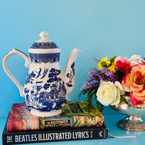 May include: A white and blue ceramic teapot with a lid, featuring a detailed landscape design. The teapot is placed on top of two books, including "The Beatles Illustrated Lyrics". A bouquet of colorful flowers sits nearby.