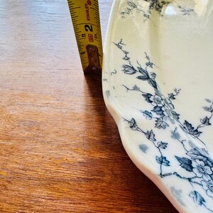 May include: A white ceramic serving platter with a blue floral design. The platter is approximately 2 inches wide, as measured by a yellow measuring tape. The design features delicate blue flowers and leaves.