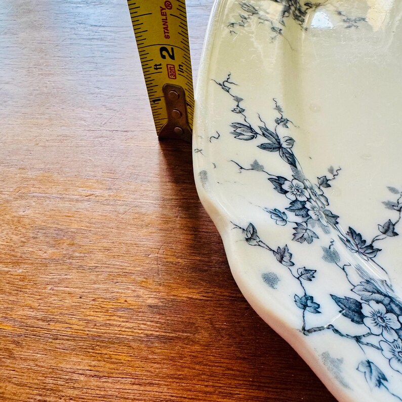 May include: A white ceramic serving platter with a blue floral design. The platter is approximately 2 inches wide, as measured by a yellow measuring tape. The design features delicate blue flowers and leaves.