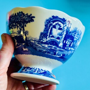 May include: A white and blue ceramic bowl with a pedestal base. The bowl features a blue transferware design of a landscape with trees, buildings, and a bridge. The rim of the bowl has a scalloped edge.
