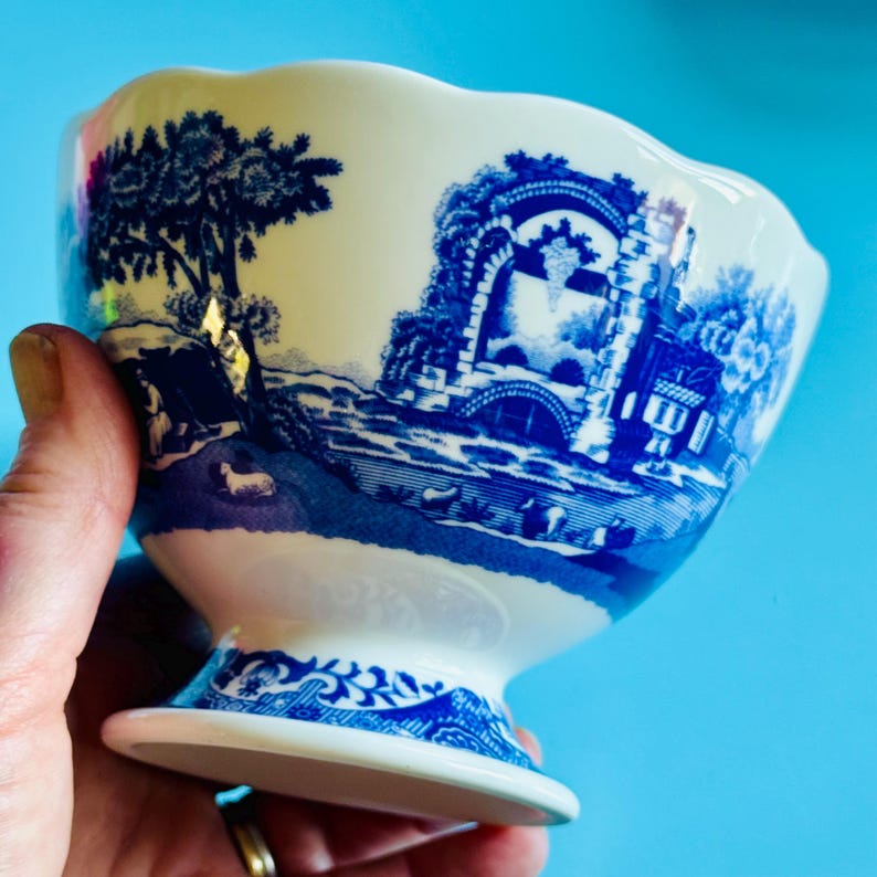 May include: A white and blue ceramic bowl with a pedestal base. The bowl features a blue transferware design of a landscape with trees, buildings, and a bridge. The rim of the bowl has a scalloped edge.