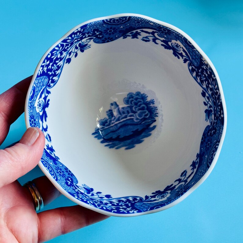 May include: A white ceramic bowl with a blue floral and landscape design. The rim has a wavy edge and is decorated with blue floral patterns. The interior features a blue landscape scene. The bowl is held against a blue background.