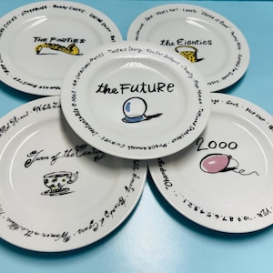 May include: A collection of white ceramic plates with black text and illustrations. Each plate features a different decade or era, including "The Forties," "The Eighties," "2000," "Turn of the Century," and "the Future."