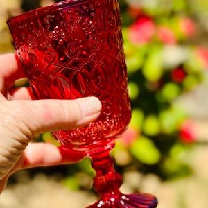 May include: A ruby red glass goblet with an ornate, embossed pattern. The stem and base are also red, and the glass is held up against a blurred background of green and red foliage. The goblet is a vintage style.