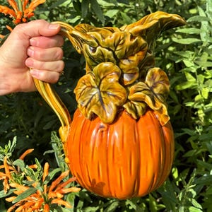 May include: A ceramic pitcher in the shape of a pumpkin, with a handle and leafy accents. The pitcher is orange with green and yellow details. The pitcher is held in front of a background of green foliage and orange flowers.