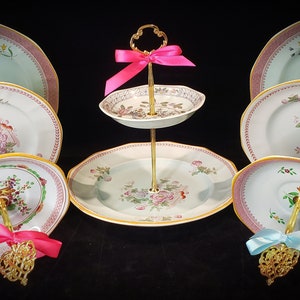 May include: A two-tiered cake stand with a gold metal base and a white porcelain top. The stand is decorated with pink and green floral patterns and has a pink and blue ribbon on each tier.