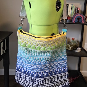 Puede incluir: Bolso de hombro de ganchillo hecho a mano con un diseño degradado, que transiciona del azul al verde al amarillo. El bolso presenta un patrón geométrico y un cierre de cremallera. La correa es de color gris claro y el bolso se muestra en un maniquí.