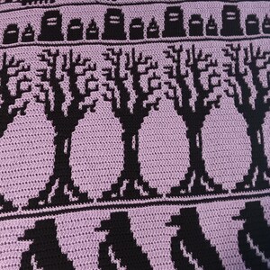 May include: Crocheted blanket with a purple background and black details. The design features a graveyard scene with a fence, tombstones, trees, and crows.