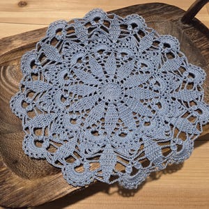 May include: A light blue, crocheted doily with a floral design. The circular doily is placed on a wooden tray. The intricate lace pattern is visible throughout the piece. The doily is a decorative item.