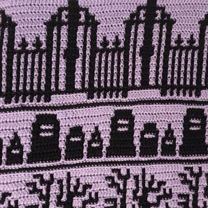 May include: A close-up of a crocheted blanket with a purple background and black details. The design features a repeating pattern of gates, tombstones, and trees, creating a spooky and whimsical Halloween theme.
