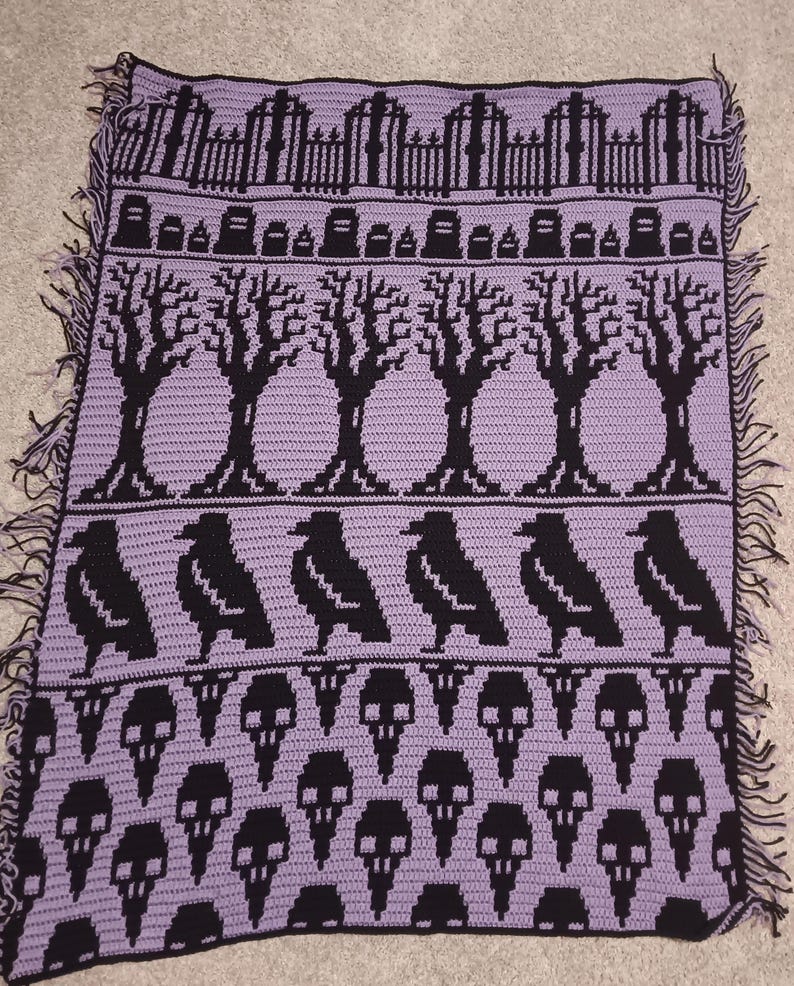 May include: A crocheted blanket with a spooky Halloween theme. The blanket features a purple background with black crocheted images of a graveyard, trees, crows, and skulls.