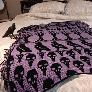 May include: A purple and black crocheted throw blanket with a pattern of trees, ravens, and skulls. The blanket has a fringe border.