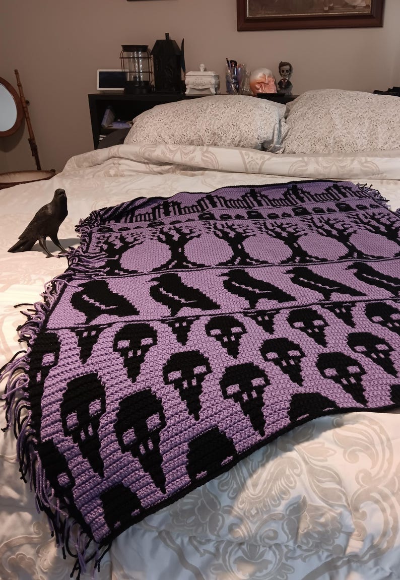 May include: A purple and black crocheted throw blanket with a pattern of trees, ravens, and skulls. The blanket has a fringe border.