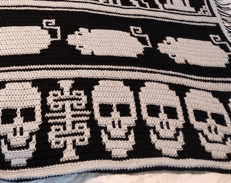 May include: Black and white crochet blanket with a repeating pattern of skulls and stylized trees. The blanket is made with a chunky yarn and has a textured look.