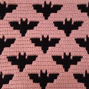 Könnte beinhalten: Eine gehäkelte Decke mit einem sich wiederholenden Muster aus schwarzen Fledermäusen auf hellrosa Hintergrund. Die Fledermäuse sind gleichmäßig verteilt und erzeugen ein strukturiertes Design zum Thema Halloween. Die Decke scheint handgefertigt zu sein.