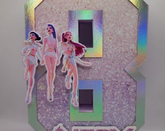 May include: A large, iridescent number eight-shaped decoration with three cartoon figures and the name "ANERY" at the bottom. The number has a holographic border and a light pink background with two rectangular cutouts.