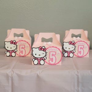 May include: Three pink party favor boxes with white bows on top. Each box features a Hello Kitty graphic and a pink circle with the number 5 and the name "Alexa is" printed on it. The boxes are on a white surface.