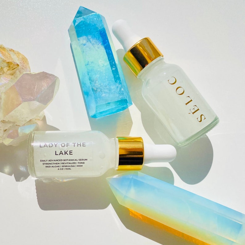 May include: Two glass serum bottles with gold accents and white droppers are displayed with crystals. One bottle reads "LADY OF THE LAKE" and the other reads "S&Eacute;LOC". The bottles are surrounded by blue and iridescent crystals.
