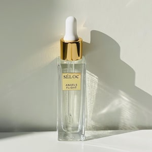 K&ouml;nnte beinhalten: Eine klare Glasflasche mit einer wei&szlig;en Pipette und einem goldfarbenen Verschluss. Die Flasche hat eine rechteckige Form und ein Etikett mit der Aufschrift "S&Eacute;LOC ANGELS FLIGHT". Bei der Flasche handelt es sich wahrscheinlich um ein Hautpflegeprodukt oder Parf&uuml;m.