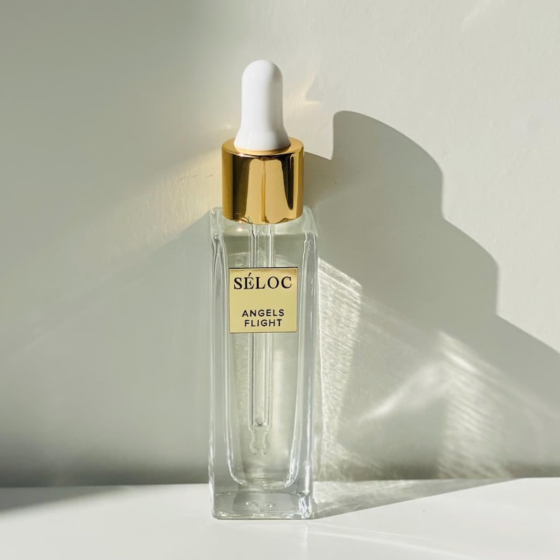 K&ouml;nnte beinhalten: Eine klare Glasflasche mit einer wei&szlig;en Pipette und einem goldfarbenen Verschluss. Die Flasche hat eine rechteckige Form und ein Etikett mit der Aufschrift "S&Eacute;LOC ANGELS FLIGHT". Bei der Flasche handelt es sich wahrscheinlich um ein Hautpflegeprodukt oder Parf&uuml;m.