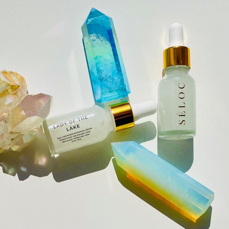 May include: Two glass bottles of serum with gold accents and white droppers, one labeled "LADY OF THE LAKE" and the other "S&Eacute;LOC". The bottles are surrounded by colorful crystal formations in shades of blue, yellow, and clear.