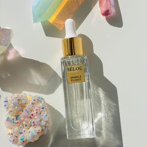 K&ouml;nnte beinhalten: Eine klare Glasflasche mit goldenem Verschluss und wei&szlig;er Pipette. Die Flasche tr&auml;gt ein Etikett mit der Aufschrift "S&Eacute;LOC ANGELS FLIGHT". Die Flasche ist von bunten Kristallen umgeben, vor wei&szlig;em Hintergrund.