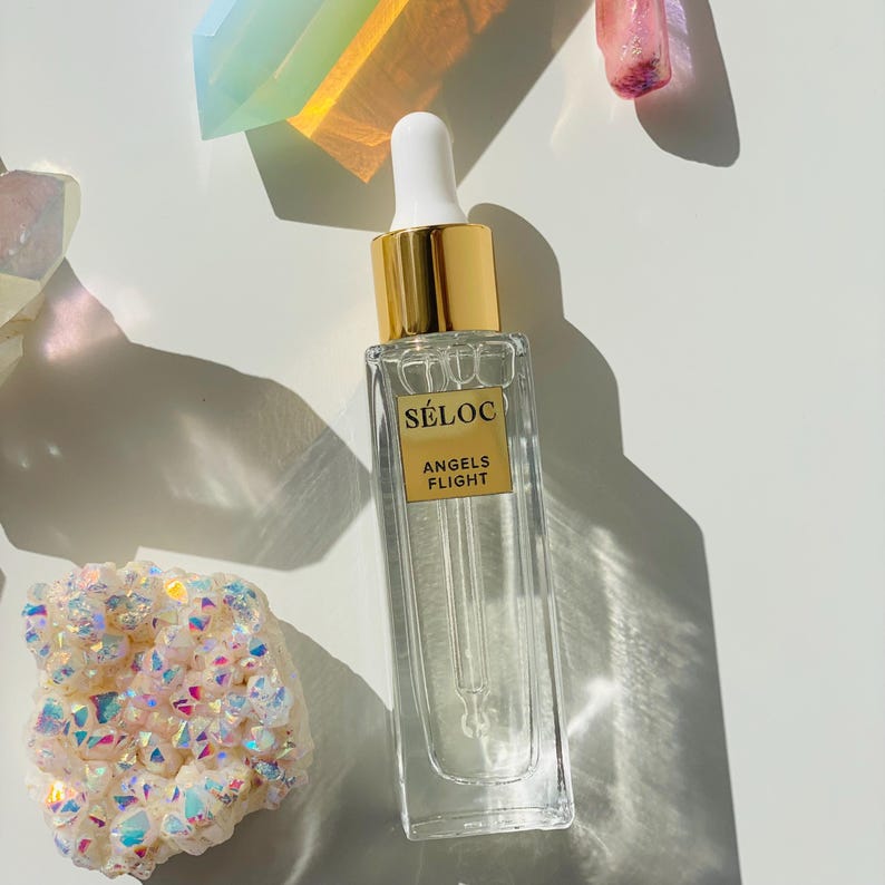 K&ouml;nnte beinhalten: Eine klare Glasflasche mit goldenem Verschluss und wei&szlig;er Pipette. Die Flasche tr&auml;gt ein Etikett mit der Aufschrift "S&Eacute;LOC ANGELS FLIGHT". Die Flasche ist von bunten Kristallen umgeben, vor wei&szlig;em Hintergrund.