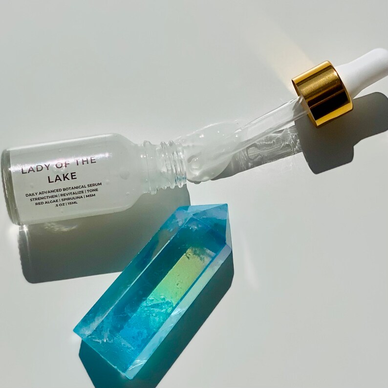 May include: A clear glass serum bottle with a gold dropper and the text "LADY OF THE LAKE" is next to a blue and green crystal. The bottle contains a clear liquid. The text on the bottle reads "DAILY ADVANCED BOTANICAL SERUM".