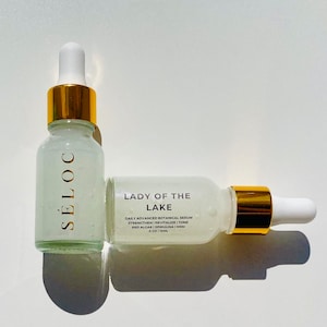 May include: Two glass serum bottles with white droppers and gold accents. One bottle is upright, the other is lying down. The bottles have the text "S&Eacute;LOC" and "LADY OF THE LAKE" with product information. The serum is 15ml.