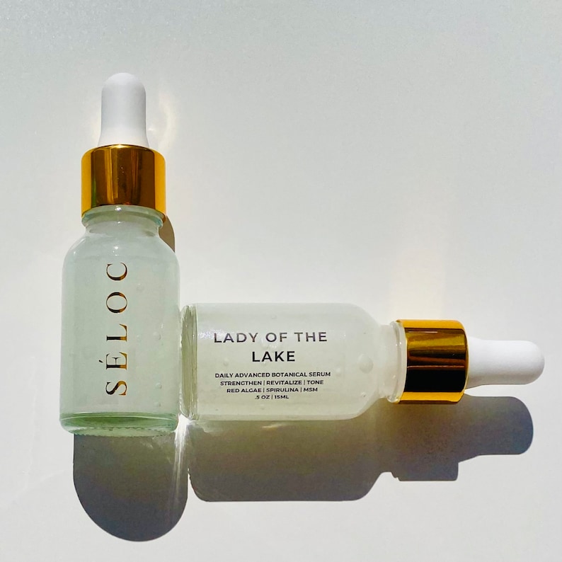 May include: Two glass serum bottles with white droppers and gold accents. One bottle is upright, the other is lying down. The bottles have the text "S&Eacute;LOC" and "LADY OF THE LAKE" with product information. The serum is 15ml.