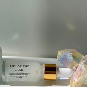 May include: A white bottle of "Lady of the Lake" daily advanced botanical serum. The bottle has a gold-colored cap and a white dropper. The label lists ingredients like red algae and spirulina. The background includes crystals.